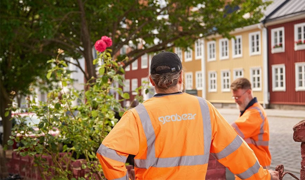 Vy över två personer i orange Geobear-kläder med en rosenbuske bredvid och färgglada hus i bakgrunden
