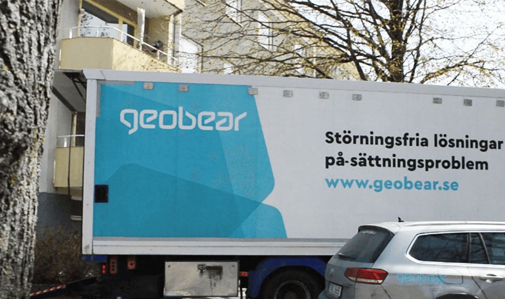 Geobears lastbil som är vit och blå står stilla framför ett lägenhetshus med en silvrig bil framför.